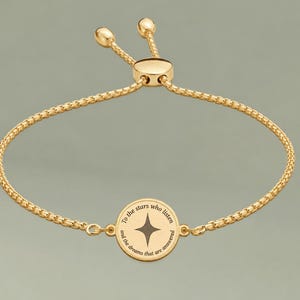 May include: A gold-toned adjustable bolo bracelet featuring a central circular charm. The charm displays a four-pointed star icon in the center, surrounded by the engraved text: To the stars who listen and the dreams that are answered.
