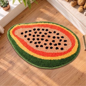 Puede incluir: Una alfombra de pelo cortado con forma de media papaya, que presenta una sección central de color naranja claro con puntos negros dispersos, rodeada de anillos concéntricos de color naranja brillante, amarillo y un borde exterior verde oscuro.