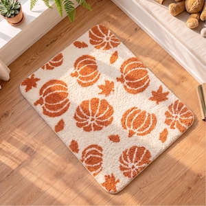 May include: A rectangular area rug with a cream-coloured background featuring a repeating pattern of orange pumpkins and scattered autumn leaves. The rug has a soft, textured pile and rounded corners, displaying a consistent seasonal motif.