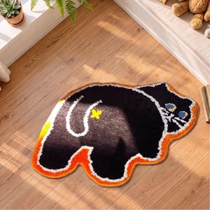 May include: A tufted rug shaped like a black cat with a white outline, blue eyes, and a yellow cross symbol on its back. The rug features a thick orange border and a textured surface, depicting a stylized feline figure in a recumbent position.