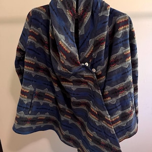 Peut inclure: Une veste à motifs bleus, gris et noirs avec un large col châle et deux boutons blancs sur le devant. Le vêtement présente un motif géométrique répétitif dans des tons de bleu, gris, noir et des touches de rouge, avec des manches longues.
