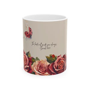 May include: A beige ceramic mug featuring a watercolor illustration of red and pink roses at the base, two ladybugs, and the black script text "The Lord will guide you always... - Isaiah 58:11" printed in the center of the mug's exterior surface.