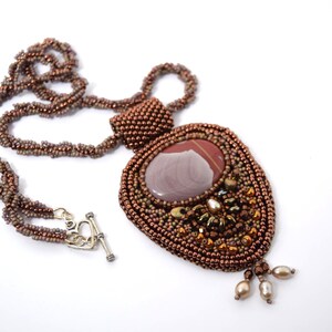 May include: A beaded necklace featuring a large, smooth, oval-shaped jasper cabochon in shades of brown and gray. The pendant is surrounded by intricate bronze-colored seed bead embroidery, accented with small pearls and a silver-toned heart toggle clasp.