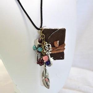May include: A brown leather journal necklace with a silver key charm, turquoise and burgundy beads, a pink heart charm, and a silver feather charm.