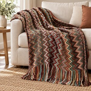 Könnte beinhalten: Eine gewebte Wohndecke mit einem mehrfarbigen Zickzackmuster in Rost, Petrol, Creme und Dunkelbraun. Die Decke hat eine strukturierte Strickoptik und ist am unteren Rand mit langen, mehrfarbigen Fransen versehen, die den Abschluss bilden.