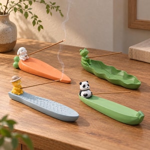 Può includere: Quattro porta incenso in ceramica con piccole statuette di animali: un coniglio bianco su una carota arancione, una rana verde su una foglia, un gatto giallo con cappello su una base blu a forma di pesce e un panda su una base verde.