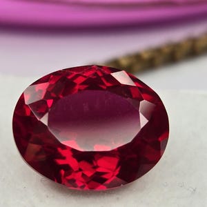 May include: A deep red, oval-cut gemstone with a faceted surface that reflects light. The stone displays a rich, saturated crimson hue and is positioned on a white surface, showcasing its transparent clarity and precise geometric cuts.