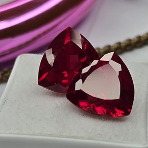 Puede incluir: Dos piedras preciosas de color rojo intenso con talla trillante y superficies facetadas. Las piedras se muestran sobre un pequeño joyero cuadrado forrado de terciopelo blanco. El color rojo vibrante es translúcido y brillante.