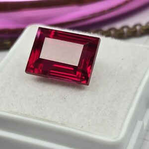 May include: A rectangular, faceted gemstone with a deep, vibrant red color. The stone is set on a white, textured display pad, showcasing its clear, polished surface and sharp, geometric edges that reflect light across its top facets.