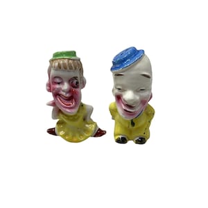 May include: Two ceramic salt and pepper shakers shaped like clown figures. One figure wears a green hat and yellow dress, winking with a wide smile. The other figure wears a blue hat and yellow suit, with a painted white face and red cheeks.