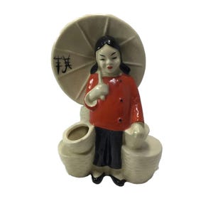 May include: A ceramic figurine of a person wearing a red top and black pants, holding a large beige umbrella with black characters. The figure stands between two woven-style baskets, one of which is open at the top, featuring a glossy finish.
