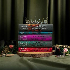 Puede incluir: Una pila de cinco libros de Sarah J. Maas titulados A Court of Silver Flames, Frost and Starlight, Wings and Ruin, Mist and Fury y Thorns and Roses, coronada con una corona de metal decorativa con piedras claras y detalles de filigrana.