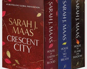 Può includere: Un cofanetto di libri intitolato Crescent City di Sarah J. Maas, contenente tre volumi: House of Earth and Blood, House of Sky and Breath e House of Flame and Shadow. Il cofanetto è rosso scuro con dettagli dorati, mentre i dorsi sono rossi, blu e neri.