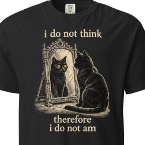 May include: A black t-shirt featuring a graphic of a black cat sitting in front of an ornate standing mirror. The cat's reflection has bright yellow eyes. The text above the mirror reads i do not think and below the mirror reads therefore i do not am.
