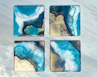Puede incluir: Juego de cuatro posavasos cuadrados con diseño abstracto. Presentan una mezcla de resina azul, blanca y dorada con líneas de acento negras y blancas. Los posavasos tienen un acabado brillante y están dispuestos sobre una superficie de mármol.