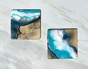 Puede incluir: Dos posavasos cuadrados con un diseño abstracto. Los posavasos presentan una mezcla de resina turquesa, blanca y dorada, con líneas onduladas negras y plateadas. El diseño se asemeja a un paisaje costero.