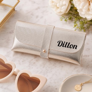 May include: A rectangular metallic gold eyeglass case with a central vertical strap and a small gold-toned snap button closure. The name Dillon is printed in black script on the front flap of the case, which has a smooth, reflective finish.