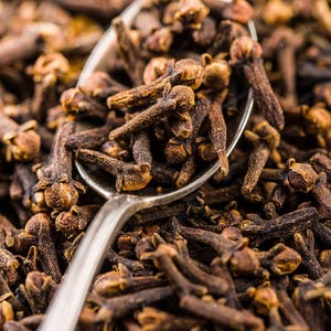 May include: A silver metal spoon filled with dried brown whole cloves, resting on a large pile of identical dried cloves. The cloves have a textured, woody appearance with a small, rounded bud at one end and a narrow, tapered stem at the other.