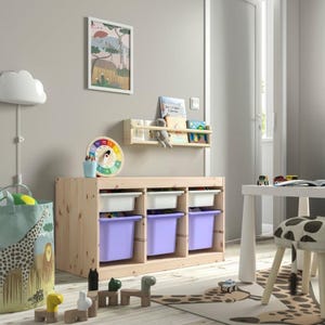 May include: A light-colored wooden toy storage unit with three white upper bins and three purple lower bins. A wooden wall-mounted shelf holds books above the unit. A fabric bin with a giraffe print and a wooden toy set are on the floor.