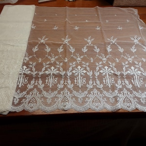 May include: A sheer white lace fabric featuring an intricate embroidered pattern of crosses and religious symbols along the scalloped bottom edge. The textile displays a repeating floral motif above the crosses, with a folded section on the left side.