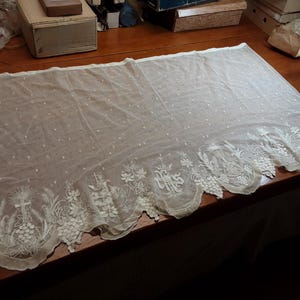 May include: A rectangular off-white lace altar cloth featuring a delicate Swiss dot pattern across the main body and an intricate scalloped border. The hem is decorated with embroidered religious motifs including grapes, wheat, and the letters IHS.