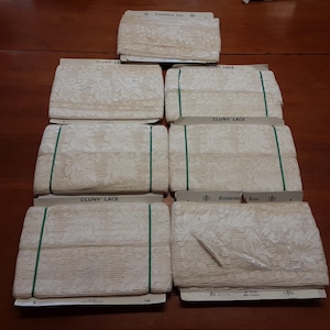 May include: Seven rolls of off-white Cluny lace trim wrapped around rectangular cardboard cards. Each roll features a repeating leaf pattern and a textured mesh background. The lace is secured with thin green elastic bands or paper labels.