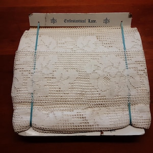 May include: A folded length of off-white ecclesiastical lace featuring a floral pattern, secured to a white cardboard header card with blue elastic bands. The header card displays the text "IHS Ecclesiastical Lace. IHS" in black ink.