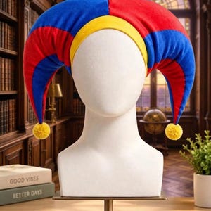 May include: A jester hat featuring a multi-coloured design with alternating red and blue fabric panels, a yellow band along the forehead, and two long, curved points that hang down on each side, each finished with a small yellow pom-pom.