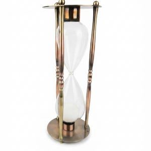 Puede incluir: Un reloj de arena con un bulbo de vidrio transparente que contiene arena blanca, sostenido dentro de un marco metálico con acabado en bronce y cobre. La estructura consta de una base y una parte superior circulares unidas por cuatro pilares.