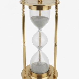 Op de afbeelding: Een zandloper met een goudkleurig metalen frame en heldere glazen bollen met grijs zand. De structuur heeft een ronde boven- en onderkant die verbonden zijn door vier verticale zuilen. De letter F is zichtbaar op de onderste glazen bol.