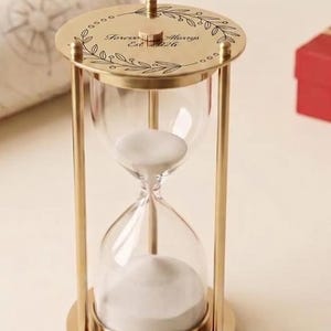 May include: A gold-toned metal hourglass with clear glass bulbs filled with white sand. The top circular plate features an engraved leaf design and the text Forever Always Est 2026. The frame consists of thin vertical pillars.