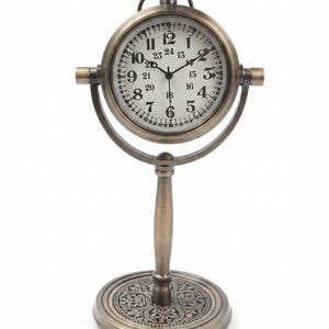 May include: A bronze-toned metal table clock with a circular face featuring black Arabic numerals for 1 through 12 and 13 through 24. The clock is mounted on a rotating gimbal frame supported by a central pedestal stand with a decorative circular base.