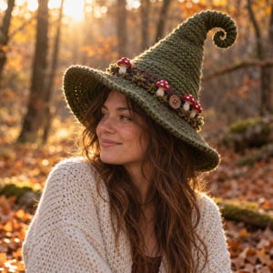 May include: A dark olive green crocheted hat with a tall, pointed, curled top. The hat features a dark brown band decorated with small, white-stemmed red mushrooms and a single round wooden button, creating a textured, handcrafted appearance.