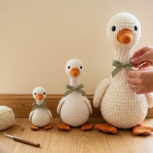 May include: Three handmade crochet geese in varying sizes, each featuring a cream-colored body, orange beak and feet, black bead eyes, and a sage green ribbon tied around the neck. The geese are arranged in a row from smallest to largest.