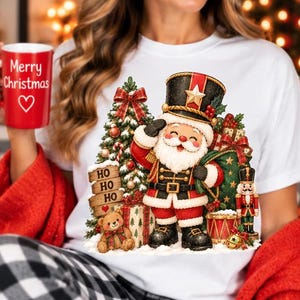 Könnte beinhalten: Ein weißes T-Shirt mit einer Grafik von einem als Nussknacker verkleideten Weihnachtsmann, neben einem Weihnachtsbaum, einem Teddybären und einem Holzschild mit der Aufschrift HO HO HO. Ein roter Becher mit Merry Christmas und Herz ist sichtbar.