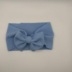 Puede incluir: Una diadema de color azul claro con un tejido texturizado tipo gofre y un lazo grande centrado. El accesorio está confeccionado con un material suave y elástico que presenta un color sólido y uniforme en toda la pieza.