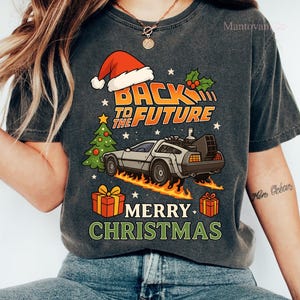 Könnte beinhalten: Ein anthrazitfarbenes T-Shirt mit einer Grafik eines silbernen Sportwagens mit Flammen, einer Weihnachtsmannmütze, einem Weihnachtsbaum und zwei Geschenken. Der Text lautet BACK TO THE FUTURE und MERRY CHRISTMAS in orange, weiß und grün.