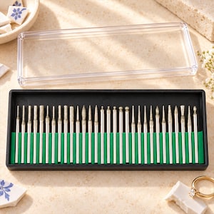 May include: A set of thirty silver-toned metal rotary tool bits with various abrasive tip shapes, including ball, needle, and cone points. The bits are organised in a black rectangular storage case with a clear lid and a green insert.