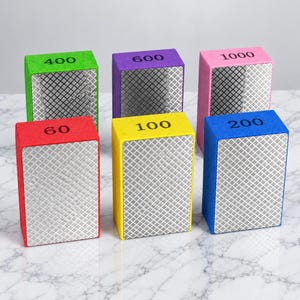 May include: Six rectangular diamond polishing blocks with foam bases in red, yellow, blue, green, purple, and pink. Each block features a metallic diamond-patterned surface and is labeled with grit numbers 60, 100, 200, 400, 600, and 1000.
