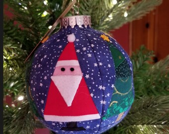 Puede incluir: Un adorno navideño de tela azul y blanco con un diseño de patchwork que presenta a Papá Noel, un árbol de Navidad y estrellas.