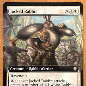 May include: A white rabbit warrior creature card from the Magic: The Gathering trading card game. The card is called "Jacked Rabbit" and has a power and toughness of 1/2. The card has the ability "Ravenous: Whenever Jacked Rabbit attacks, create a number of 1/1 white Rabbit creature tokens equal to Jacked Rabbit's power."