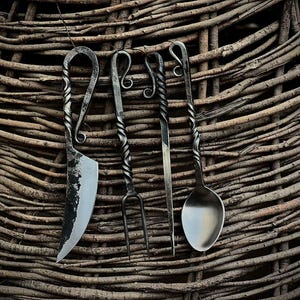 May include: A set of four hand-forged metal utensils featuring twisted handles and curled ends. The set includes a curved blade knife, a two-pronged fork, a thin pointed skewer, and a rounded spoon, all displaying a dark, textured finish.