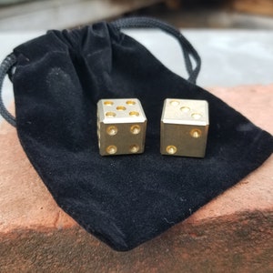 May include: Two gold metal dice with black dots on each side, resting on a black velvet pouch with a drawstring.