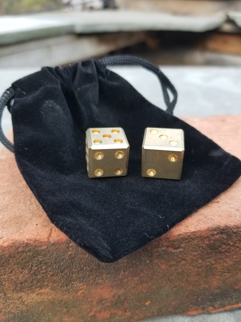 May include: Two gold metal dice with black dots on each side, resting on a black velvet pouch with a drawstring.