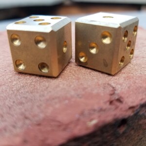 May include: Two gold metal dice with rounded edges, sitting on a red surface.