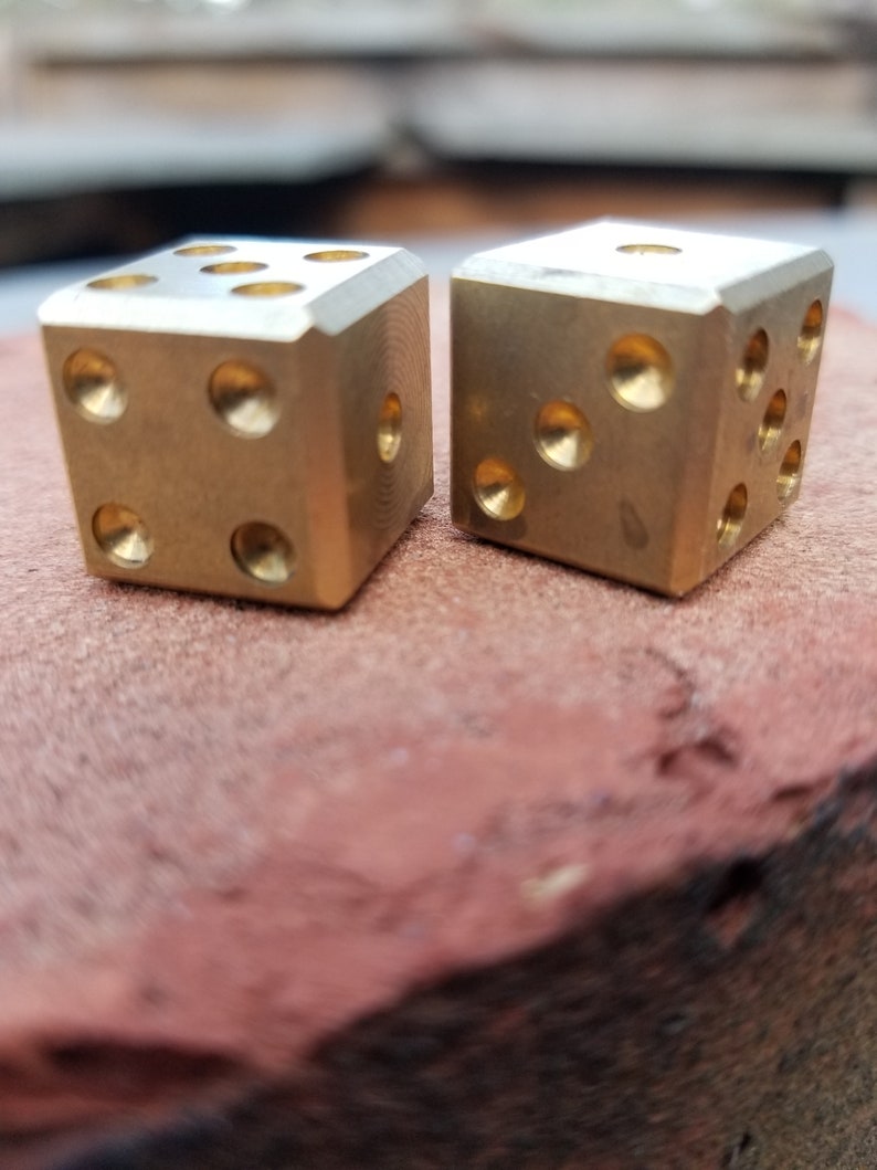 May include: Two gold metal dice with rounded edges, sitting on a red surface.