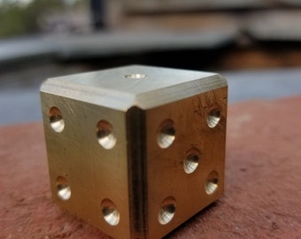 May include: A gold metal dice with rounded edges and six sides. Each side has a different number of dots.