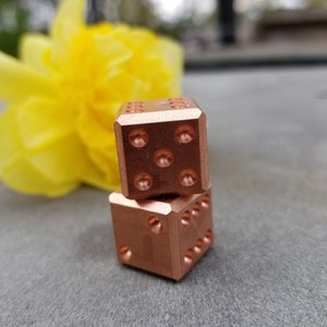 May include: Two copper dice stacked on top of each other. The dice are a dark copper colour and have a smooth, polished finish.