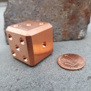 May include: A copper-colored metal dice with rounded edges and six sides. The dice is sitting on a gray, textured surface next to a copper-colored US penny.