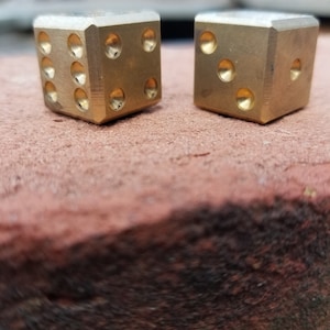 May include: Two gold metal dice with rounded edges. The dice are sitting on a brown surface.
